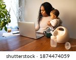 A beautiful young mixed Race African American mother holds her daughter while taking notes at her dining table serving as a temporary remote work from home station with breast pump in foreground.