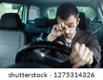 Sleepy young man rubs his eyes with his right hand. His left hand is on the steering wheel. He is sitting at his car. Road safety concept.