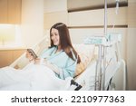 Small photo of patient woman browsing smartphone while sitting on bed in recovery room of hospital, clinic, giving saline, Asian Patient woman sick saline intake.