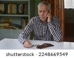 Small photo of Sad old man sits at the table, looks at the money and wallet. The concept of poverty, low income, austerity in old age.