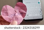 Small photo of A broken heart victim holding a crumpled heart with the words Love Scam written. Off focus background with a mobile phone message to the scammer