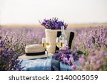 Small photo of Two glasses with white wine and bottle on background of a lavender field. Straw hat and basket with flowers lavender on a blanket on picnic. Romantic evening in sunset rays. Summer in Provence, France