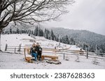 Couple in love with blanket hugs in winter on hill near wood forest. Man and woman holding cup drinking coffee. Recreation area, fire pit, fireplace in backyard house. Cabin terrace with mountain view