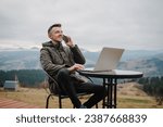 Workplace in country on backyard house in morning. Man talking on phone outdoors. Male work on a laptop sitting table with great view mountains. Concept remote work or freelance lifestyle. Internet 5G