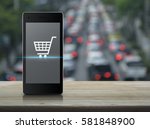 Shopping cart icon on modern smart phone screen on wooden table over blur of rush hour with cars and road, Shop online concept