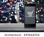 Shopping cart icon on modern smart phone screen on wooden table over blurred colourful night light city with cars, Shop online concept