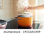 Small photo of Female hand open lid of enamel steel cooking pan on electric hob with boiling water or soup and scenic vapor steam backlit by warm sunlight at kitchen. Kitchenware utensil and tool at home background
