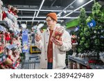 A man in a supermarket chooses Christmas tree balls and decorations. A stylish guy in a white fur coat looks at Christmas decorations on the shelves in the store, festive mood, buying Christmas gifts