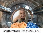 Small photo of Young blonde woman looks at the laundry in the washing machine and holds her nose against an unpleasant smell, photo from the inside