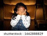 Small photo of Cute little girls sit and watch fun movies in a thrilling cinema with horror and thrilling, using hands to cover their faces, leaving only the eyes to want to thrill.