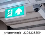 Small photo of Fire exit sign with closed-circuit television (CCTV) camera installed on ceiling structure in the building. Security and safety system for public building