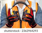 Small photo of Male worker with reflective orange vest and protective hand gloves holding yellow safety ear muffs or ear protectors preparing to wear on his head. Equipment for high noise reduction