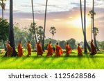 Buddhist monk and Buddhist novice with good spiritual going about with alms bowl to receive food from people in morning by walking in row across rice field with palm trees to village in Thailand