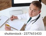 Small photo of Male medicine doctor hand hold jar of pills and write prescription to patient at worktable. Panacea and life save prescribing treatment legal drug store concept. Empty form ready to be used