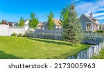 Small photo of Panorama white clouds White picket fence of a yard with green lawn and pine tree at Daybreak, Utah. There are fenced houses with large yards