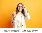Office Style. business woman in a white suit holds paper cup of coffee and puts on glasses on yellow isolated background, girl manager in jacket with drink smiles