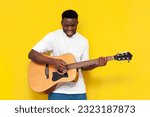 joyful african american man holds acoustic guitar and sings on yellow isolated background, the guy the artist plays musical instrument