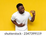 Small photo of unhappy african american man in white t-shirt eats spoiled fast food and holding on to his stomach on yellow isolated background, young guy shows stomach pain and nausea from bad burger