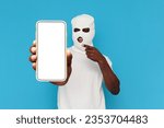 Small photo of african american man thug in white balaclava shows empty smartphone screen on blue isolated background, hoodlum in mask advertises copy space on phone