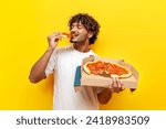 young indian man eating delicious pizza on yellow isolated background, curly guy student holding pizza box and biting a piece of fast food
