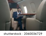 Small photo of Handsome pensive middle eastern businessman talking on mobile phone, holding financial report working with documents sitting in plane. Confident entrepreneur flying private jet, successful business