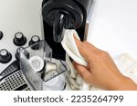 Female hand using white kitchen towel cleaning an automatic espresso machine with used coffee capsules in the blurred background.