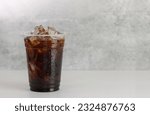 Small photo of Ice americano coffee in a tall disposable glass over grey background on white table. Cold summer drink on dramatic grey background with copy space.