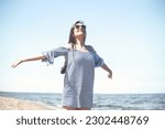Happy smiling woman in free bliss on ocean beach standing with open hands. Portrait of a brunette female model in summer dress enjoying nature during travel holidays vacation outdoors
