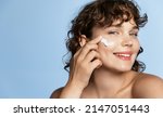 Small photo of Close up portrait of smiling young woman with curly hair, using facial skincare cream on her face, doing cosmetic daily routine after shower, standing over blue background