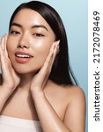Small photo of Beautiful girl with bare shoulders applying cream on her face and smiling against light-blue background. Smiling asian woman with glowing skin applying facial skincare cream