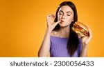 Small photo of Brunette girl licks her finger, eats tasty hamburger. Woman orders takeaway food with delivery app, enjoys delicious burger, orange background. Restaurants and takeout concept