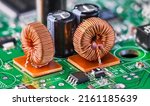 Small photo of Closeup of toroidal inductors and electrolytic capacitors on electronic PCB detail with bokeh. Two ferrite core coils with copper wire winding and various surface-mount devices on green circuit board.