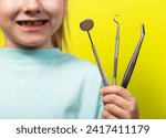 Small photo of A beautiful little girl aged seven years old holds dental instruments on a yellow background. The concept of treatment of periodontal diseases, intraosseous implantation with subsequent prosthetics.