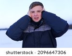 Small photo of Portrait of handsome guy, young frozen man suffering freezing because of cold ice icy weather, covering his ears, head with hands, trembling, shaking outdoors. Low temperature, coldly north weather