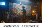 Small photo of Talented Female Artist Working on a Modern Abstract Oil Painting, Gesturing with Broad Strokes Using Paint Brush. Dark Creative Studio Large Picture Stands on Easel Illuminated, Tools Everywhere