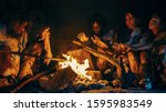 Neanderthal or Homo Sapiens Family Cooking Animal Meat over Bonfire and then Eating it. Tribe of Prehistoric Hunter-Gatherers Wearing Animal Skins Eating in a Dark Scary Cave at Night