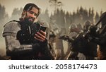 Small photo of Portrait of Handsome Medieval Knight Using Smartphone on Battlefield, Smiling. Fun Concept: Successful Armored Warrior Ordering Online, Betting, Investing, doing E-commerce. War is Raging
