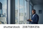 Small photo of Thoughtful Black Businessman in a Tailored Suit Using Laptop while Standing in Office Near Window on Big City. Successful Corporate Top Manager Doing Data Analysis for e-Commerce Startup