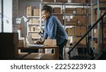 Small photo of Warehouse Female Inventory Manager Using Laptop Computer, Preparing a Small Parcel for Postage. Black Multiethnic Small Business Owner Working in Storeroom, Preparing Order for Client.