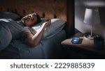 Small photo of Caucasian Man Sleeps through Smartphone Alarm Clock Showing Eight in the Morning. Tired Person Oversleeping. Person and Mobile Phone Close-up. Bedside Nightstand Bedroom Apartment