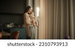 Small photo of Beautiful Young Asian Woman Holding her Baby in her Arms While Standing Next to a Window at Home. Cute Little Toddler Resting in His Mother's Embrace as She Shows him the Busy Streets of Urban City.