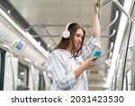 Young girl listen music in headphones and hold smartphone in subway car travel underground use wireless internet connection. Happy female chatting online in mobile phone messenger watch video in metro