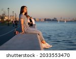 Teen skater girl sit at sidewalk during sunset at seaside. Cute young woman enjoy summer evening at river or sea looking at sun setting in water. Urban female with longboard relax alone at riverside