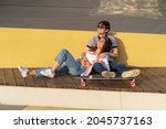 Couple in love enjoy sunset sitting embracing on longboard. Trendy urban man and woman wearing stylish sunglasses relax chilling outdoors. Happy longboarders recreate together enjoy summertime in city