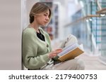 Small photo of Lover of books. Focused pensive middle-aged female passionate reader sitting on floor with book in hands, mature woman bibliophile reading in public library, soft focus. Self education