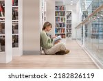 Pensive middle-aged Scandinavian woman book lover spending free leisure time in library, mature female with book in hands sitting in lotus pose on floor near bookcase, enjoying reading. Hobby concept