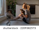 Young woman holds cat in front of electric fan and tries to cool down from stuffiness in apartment. Excited female sitting near fan helps pet to feel better from summer heat in flat kitchen