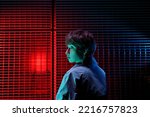 Small photo of Restrained Caucasian woman turning head to side stands with back to camera next to wall covered with mesh grating. Brown-haired girl is dressed in autumn coat on background of red blue neon glow