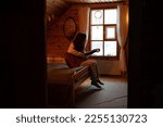 Small photo of Young woman plays guitar and enjoys music alone, sitting by window of village house with cozy bed, wooden walls. Female in warm home clothes croon melody on musical instrument while rest on vacation.