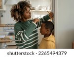Small photo of Smiling loving African American mother measuring height of child son at home, using metal ruler and pencil, marking top of head on wall, mom taking measurement while boy standing against flat surface